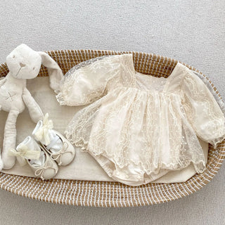 Baby outfit with lace dress, shoes, and teddy bear in a woven basket on a light background